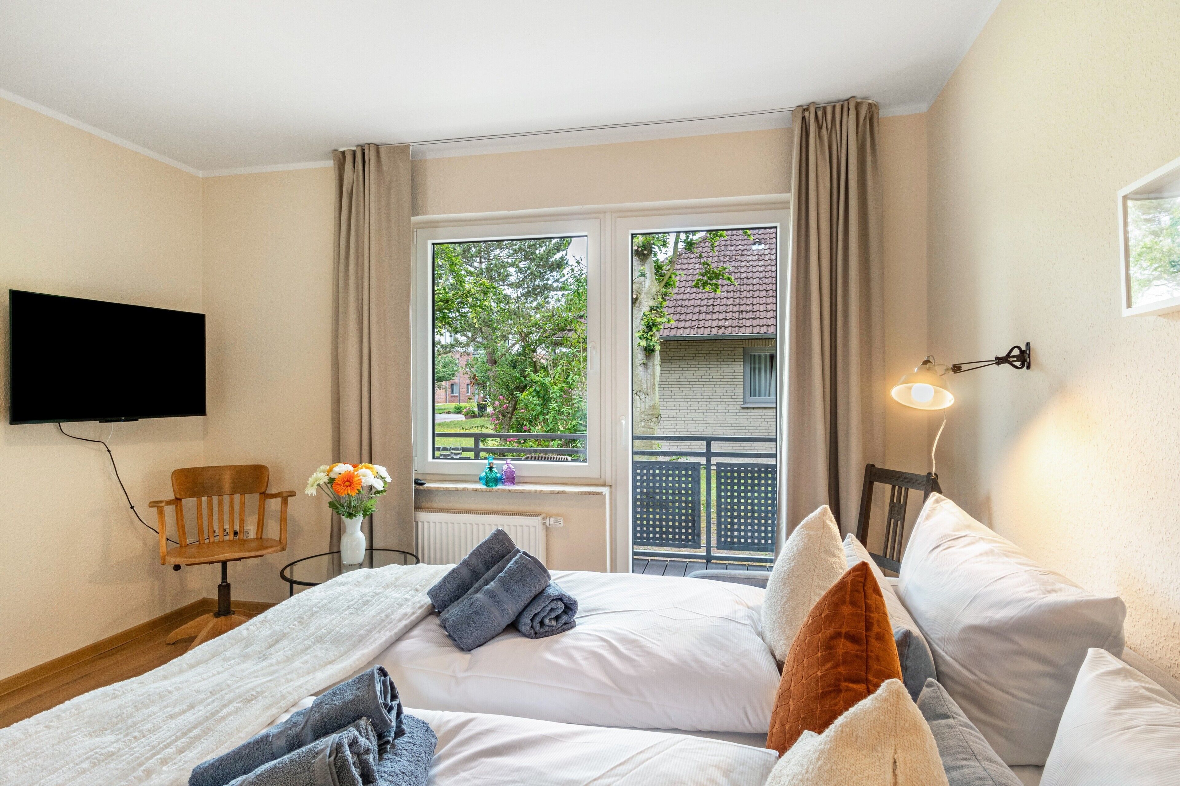 A hotel room with two beds, a TV, a window with a view of a building and trees, and a small table with a vase of flowers.