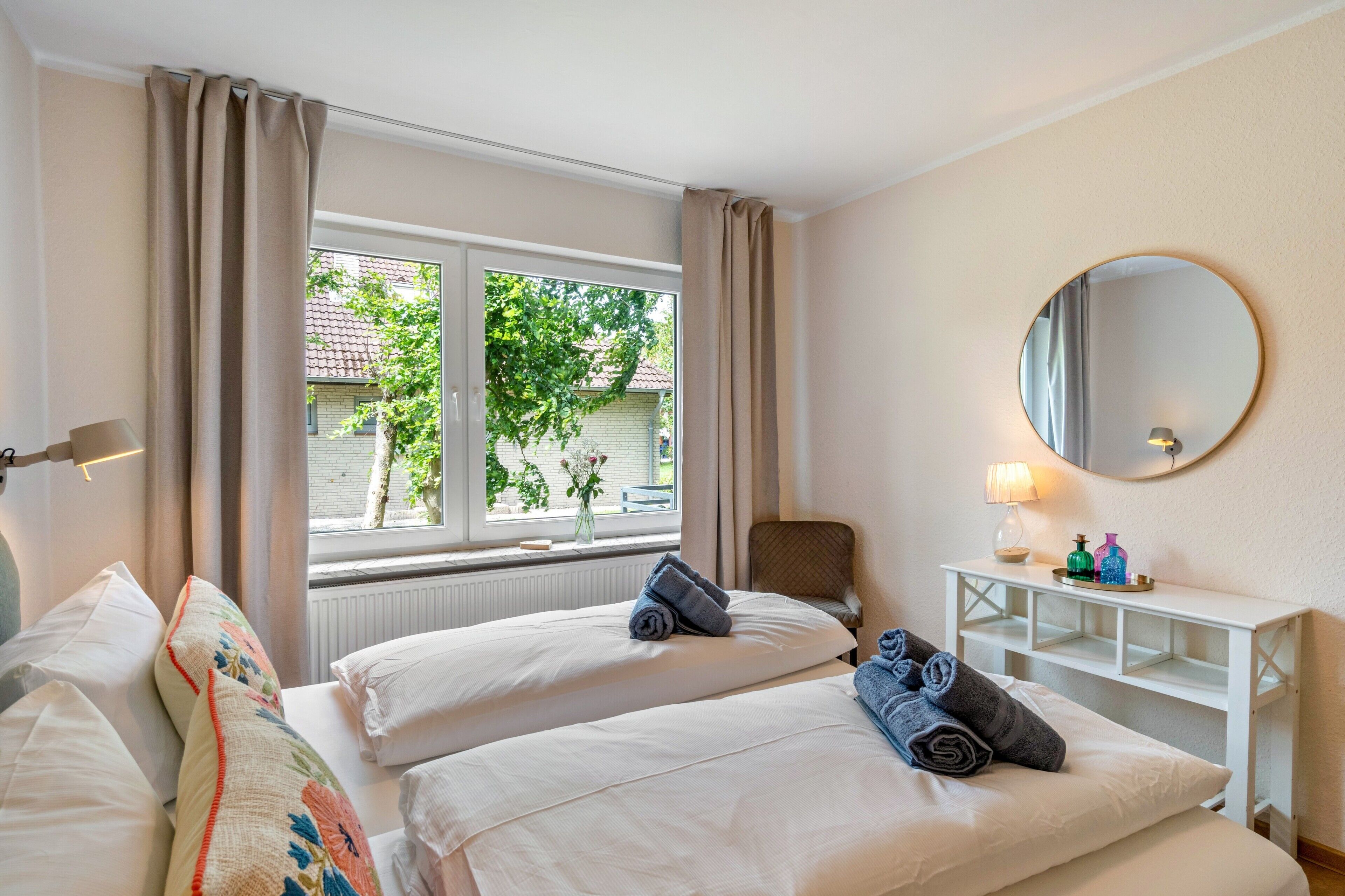 Ein Hotelzimmer mit zwei Betten, einem Fenster mit Vorhängen, einem runden Spiegel, einem Nachttisch mit Lampe und Blick auf Grünfläche.