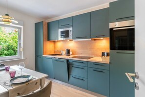 A modern kitchen with teal cabinets, stainless steel appliances, and a window with a view of greenery. - Inselheimat Apartment with Balcony (Borkum)