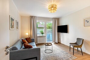 A modern living room with a grey sofa, a round coffee table, a patterned rug, a television mounted on the wall, and a window with curtains. - Inselheimat Apartment with Balcony (Borkum)