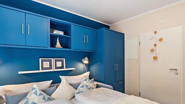 Ein Schlafzimmer mit einem Bett, blauen Schränken, einer weißen Tür und einem Regal mit Büchern und einem kleinen Schiffmodell.