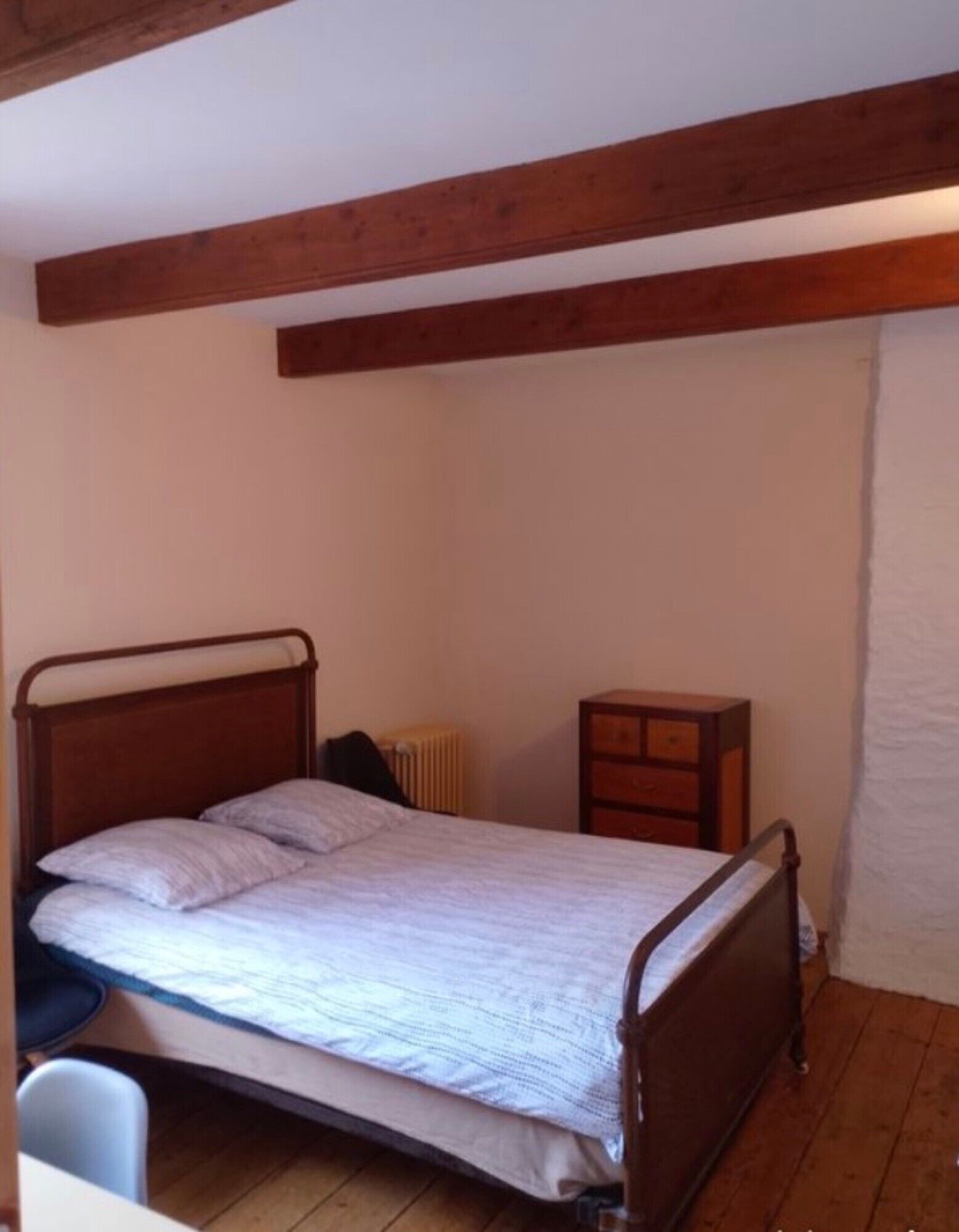 Una cama individual con estructura metálica, una mesita de noche y un radiador en una habitación con vigas de madera en el techo.
