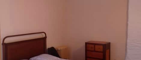 Un lit simple avec un cadre en métal, une table de chevet et un radiateur dans une pièce dont le plafond est orné de poutres en bois.