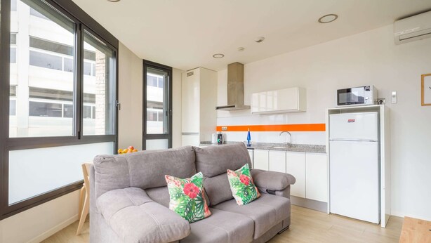 A modern living room with a grey sofa, two floral cushions, a kitchen area with white cabinets, a microwave, and a refrigerator. - Apartment in Las Palmas Near Playa de Las Canteras (Las Palmas de Gran Canaria)