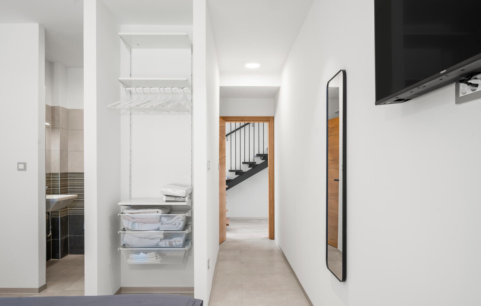 A modern interior with a hallway leading to a staircase, a white wall-mounted TV, and a metal rack with folded towels.