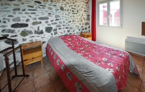 Ein Schlafzimmer mit Steinwand, einem Bett mit floralem Überwurf, einem Holfnachtkästchen, einem Heizkörper und einem Fenster mit roten Vorhängen.