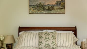 A bed with striped bedding and a floral pillow, a lamp, a vase with flowers, and a framed landscape painting on the wall.