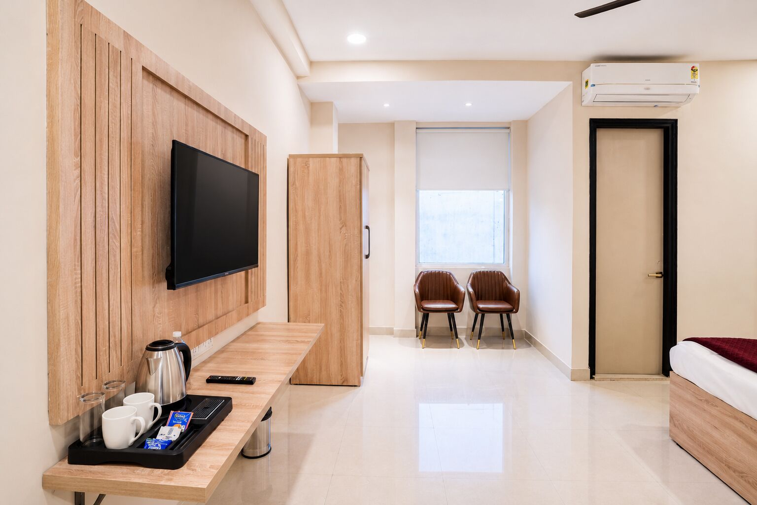 A hotel room with a TV mounted on the wall, a wooden desk with a coffee maker, two chairs, a bed, and an air conditioning unit.