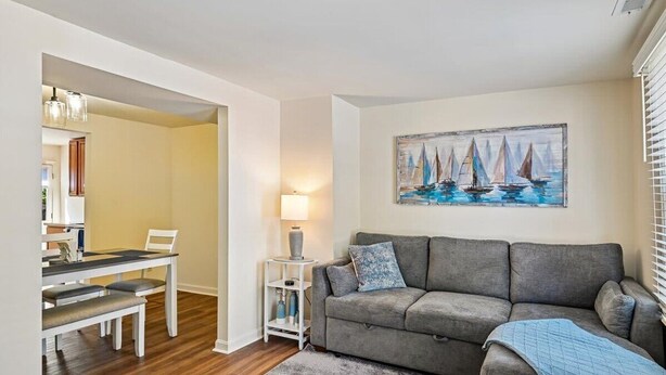 Uma sala de estar com um sofá cinzento, um quadro de barcos à vela, uma mesa de jantar com cadeiras e um abajur.