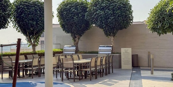 Un área de comedor al aire libre con mesas y sillas, rodeada de árboles y un muro.