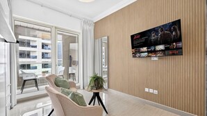 Quarto de hotel moderno com uma televisão grande na parede, área de estar com cadeiras, uma pequena mesa com uma planta e vista para a cidade pela janela.