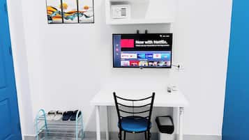 A room with a white desk, a blue chair, a TV mounted on the wall, a shelf with a microwave, and a pair of slippers on a rack.