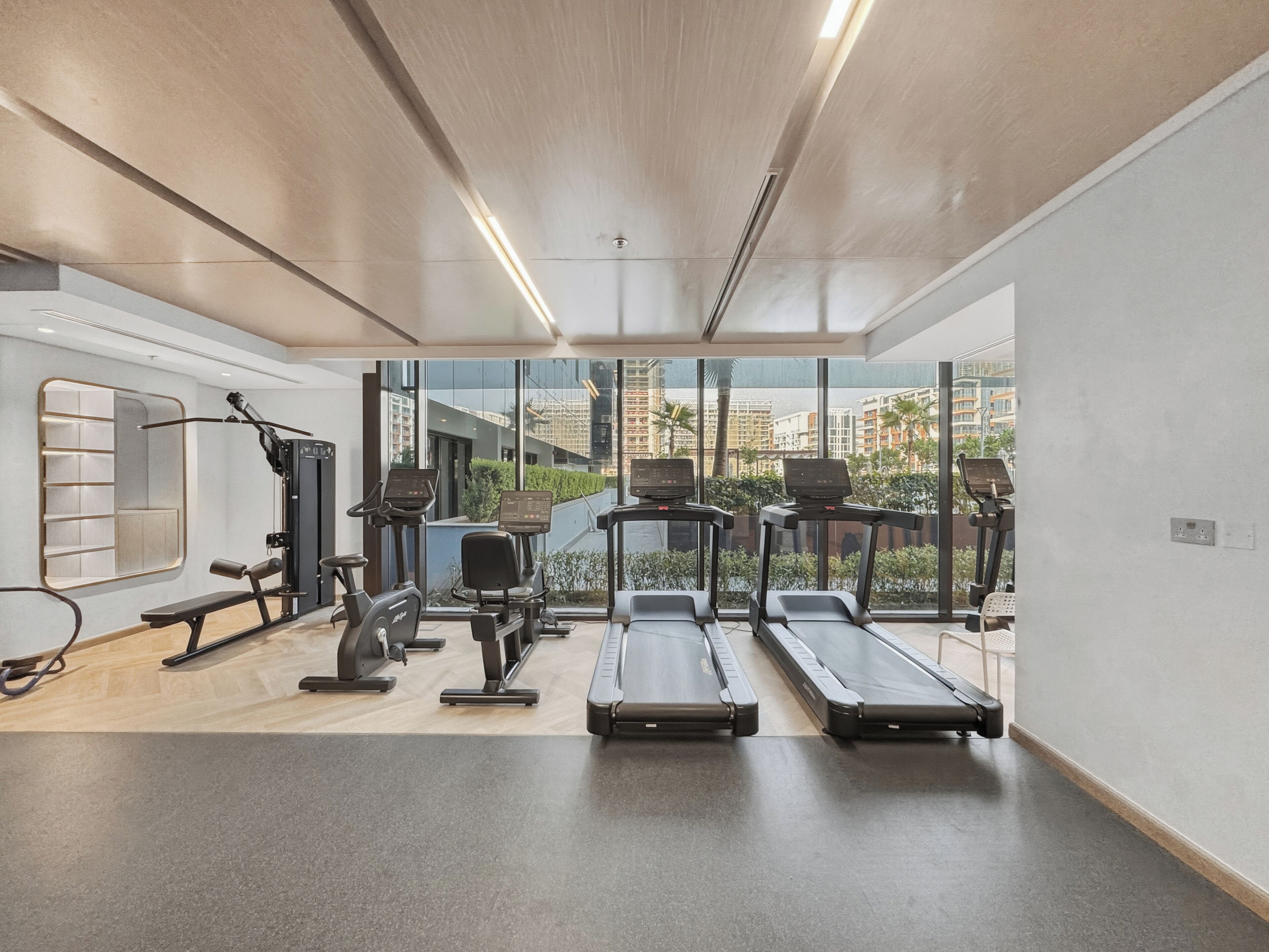 A modern gym with treadmills, elliptical machines, and a bench. Large windows offer views of buildings and greenery.