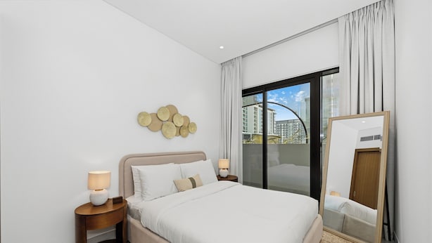 A modern hotel room with a large bed, a nightstand with a lamp, a wall-mounted decorative item, a sliding glass door, and a mirror. - Bloomfields 2BR in Azizi Riviera 51  (Dubai)