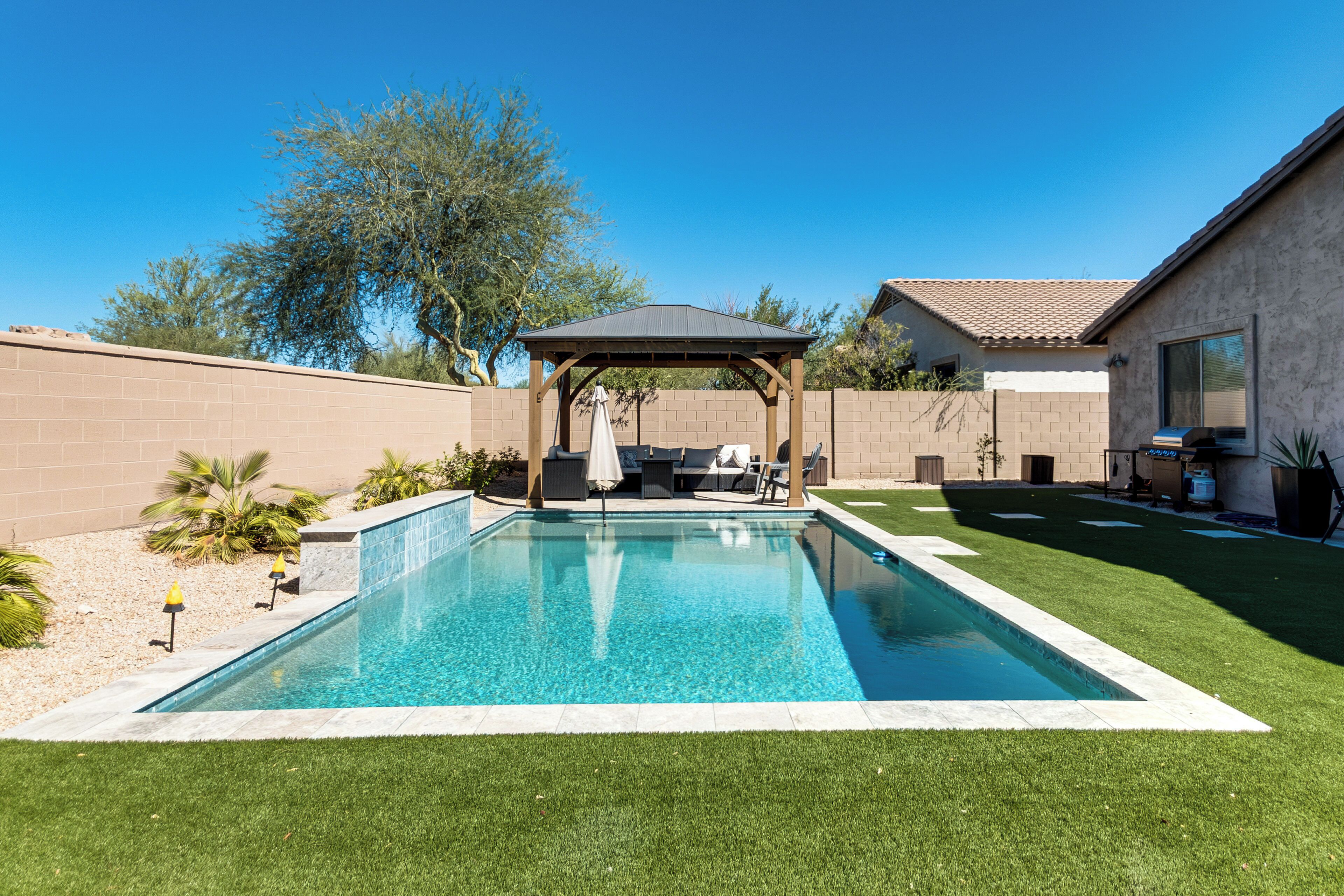 A backyard with a swimming pool, a covered seating area, and a patio with outdoor furniture.