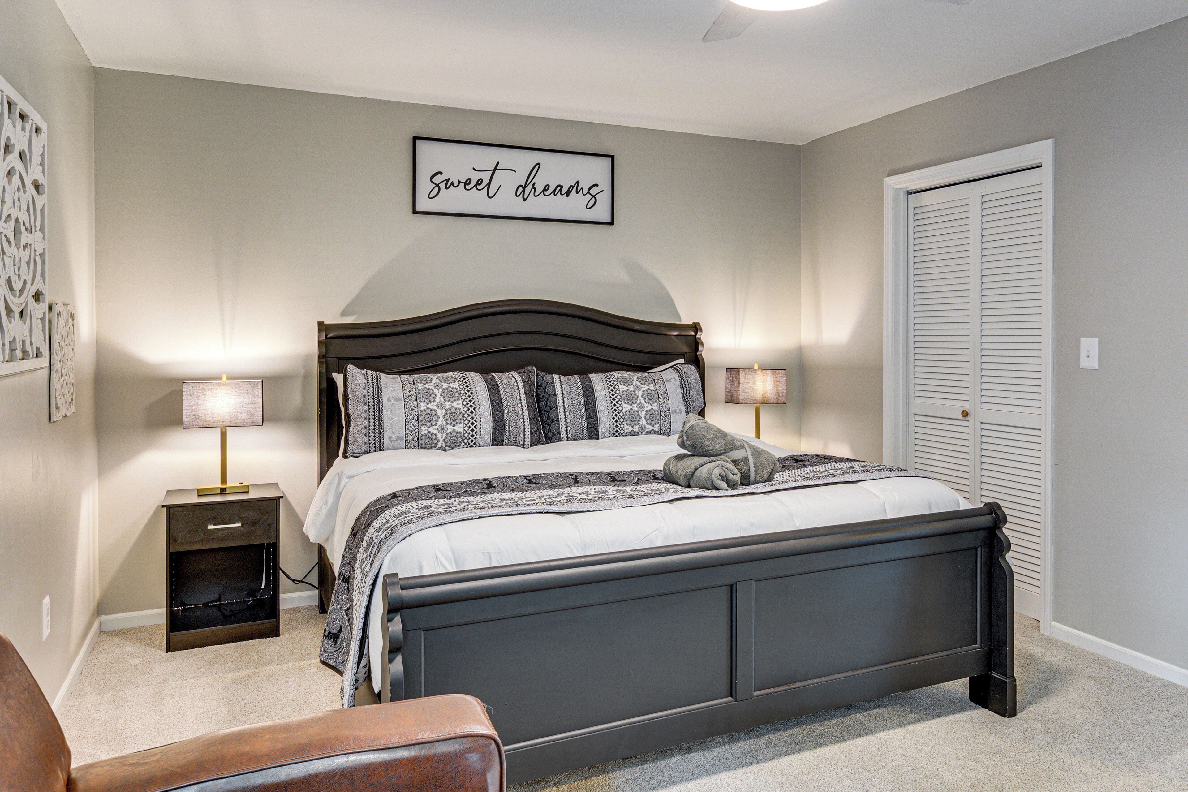 A bedroom with a bed, two bedside tables, a nightstand, a chair, a wall-mounted artwork, and a door with a window.