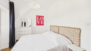A bedroom with a bed, a dresser, a small table, a wicker headboard, and a 'LOVE' sign on the wall. - Apartment for 4 - Shared Garden - Terrace - WiFi (Fouesnant)