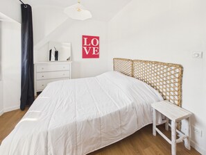A bedroom with a bed, a dresser, a small table, a wicker headboard, and a 'LOVE' sign on the wall. - Apartment for 4 - Shared Garden - Terrace - WiFi (Fouesnant)