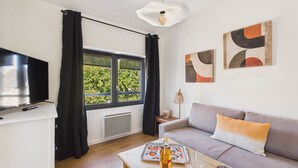 A modern living room with a sofa, a coffee table with glasses and a bottle, a TV, and a window with curtains. - Apartment for 4 - Shared Garden - Terrace - WiFi (Fouesnant)