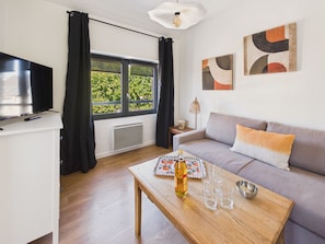 A modern living room with a sofa, a coffee table with glasses and a bottle, a TV, and a window with curtains. - Apartment for 4 - Shared Garden - Terrace - WiFi (Fouesnant)