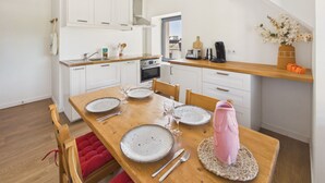 A modern kitchen with white cabinets, a wooden dining table with plates, glasses, and cutlery, and a red cushioned bench. - Apartment for 4 - Shared Garden - Terrace - WiFi (Fouesnant)