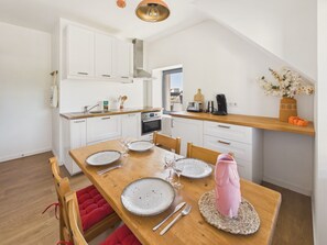 A modern kitchen with white cabinets, a wooden dining table with plates, glasses, and cutlery, and a red cushioned bench. - Apartment for 4 - Shared Garden - Terrace - WiFi (Fouesnant)
