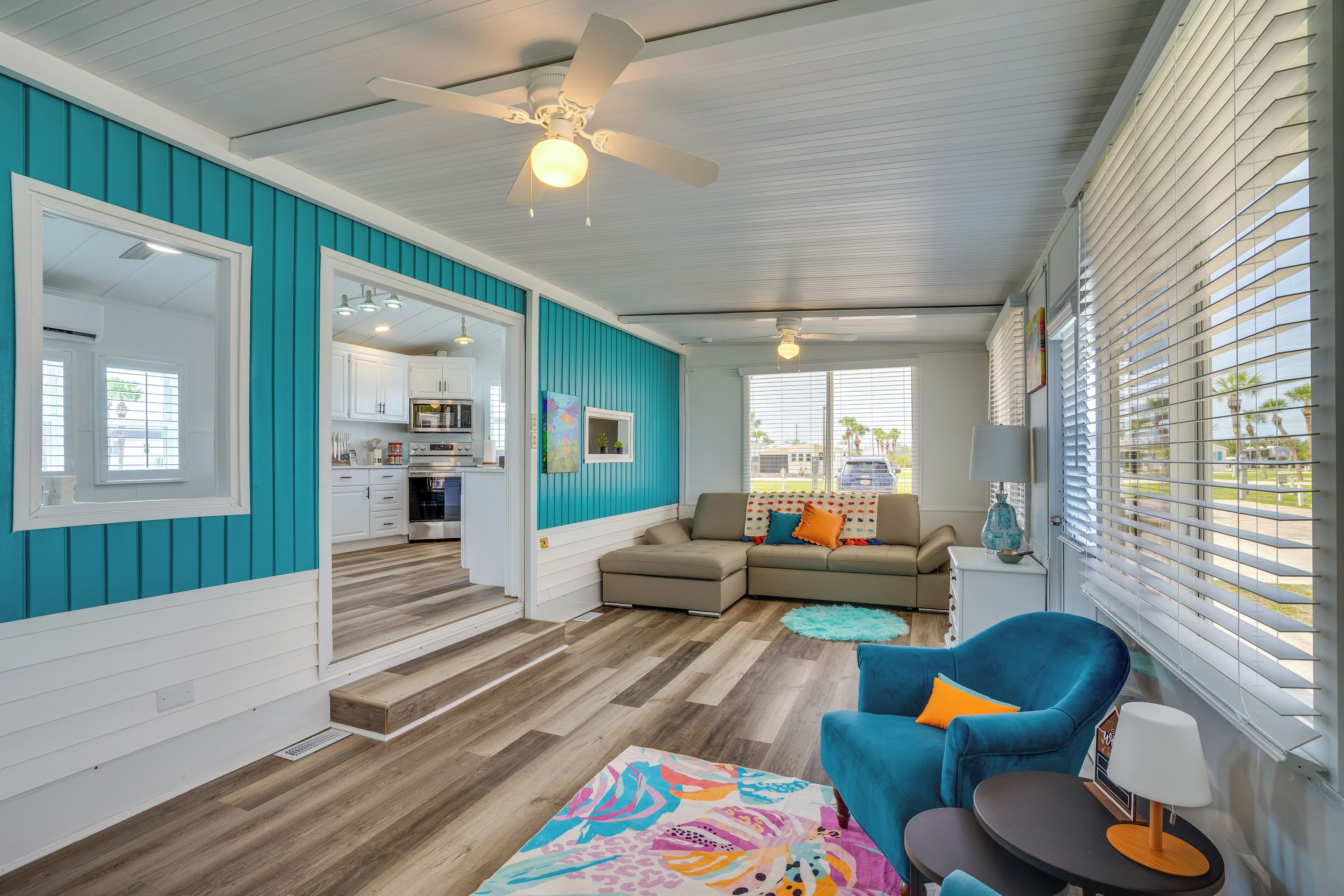 A modern living room with a teal accent wall, a ceiling fan, a sofa, a blue armchair, and a dining area visible through an open door.