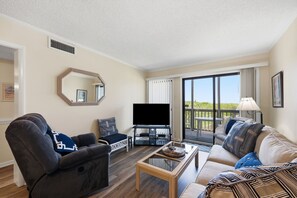 A living room with a sofa, armchair, TV, and a balcony with a view. - Dunescape 132-Ocean Front Condo w/Pool (Atlantic Beach)