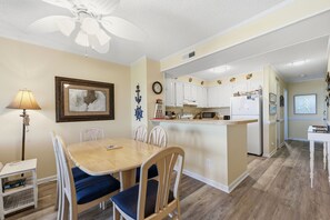 A dining area with a table and chairs, a kitchen with white cabinets, a clock, and a ceiling fan. - Dunescape 132-Ocean Front Condo w/Pool (Atlantic Beach)