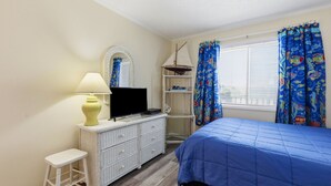 A bedroom with a bed, a television on a dresser, a lamp, a shelf with books, and a window with curtains. - Dunescape 132-Ocean Front Condo w/Pool (Atlantic Beach)