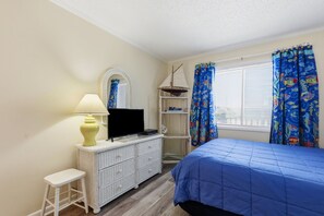 A bedroom with a bed, a television on a dresser, a lamp, a shelf with books, and a window with curtains. - Dunescape 132-Ocean Front Condo w/Pool (Atlantic Beach)
