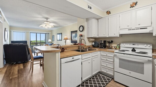 A kitchen with white cabinets, a stove, a dishwasher, and a dining area with a chair and table. - Dunescape 132-Ocean Front Condo w/Pool (Atlantic Beach)