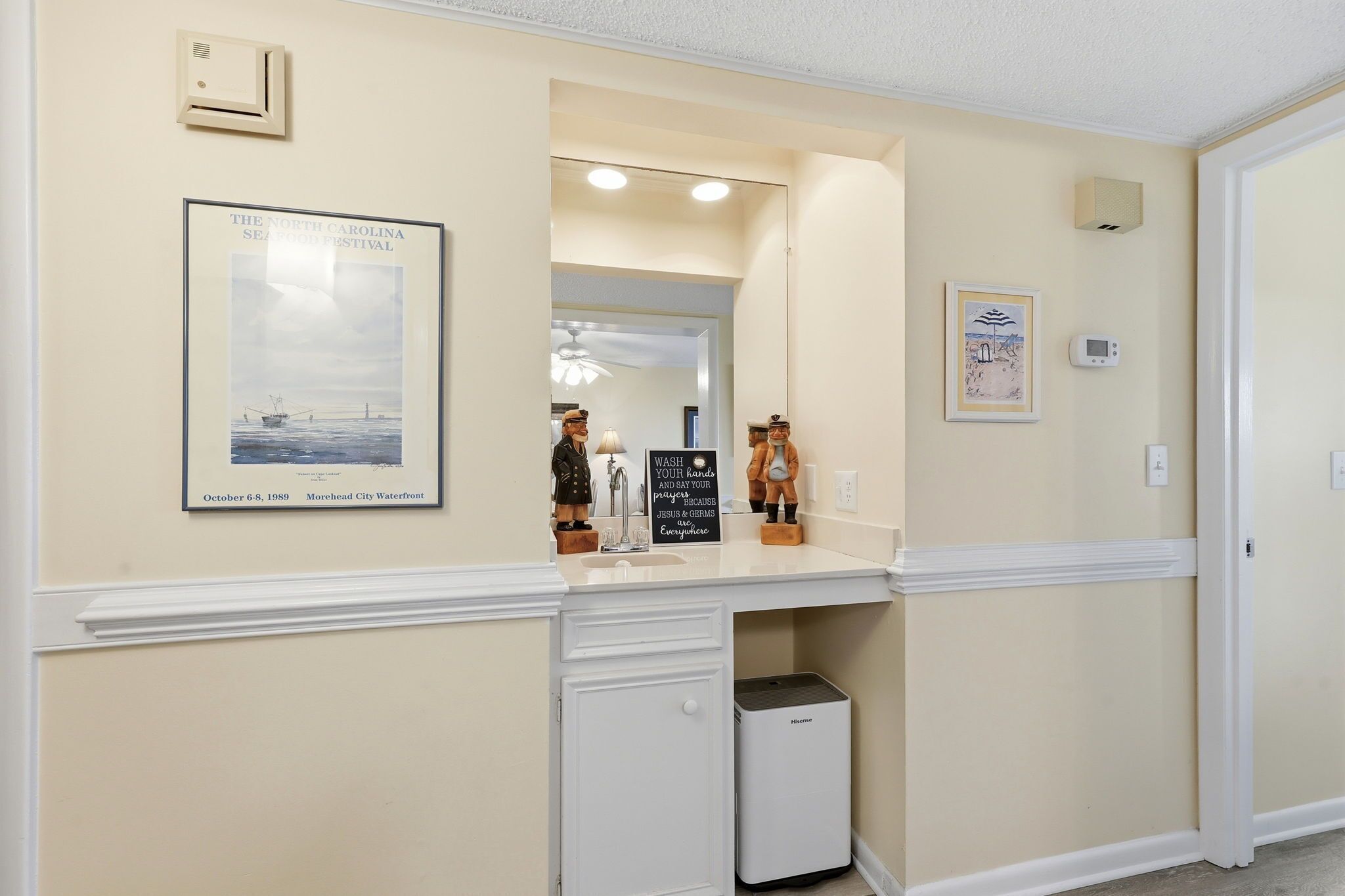 A hotel room with a white vanity, a framed poster of the North Carolina Sea Festival, a small trash bin, and a mirror reflecting the room.