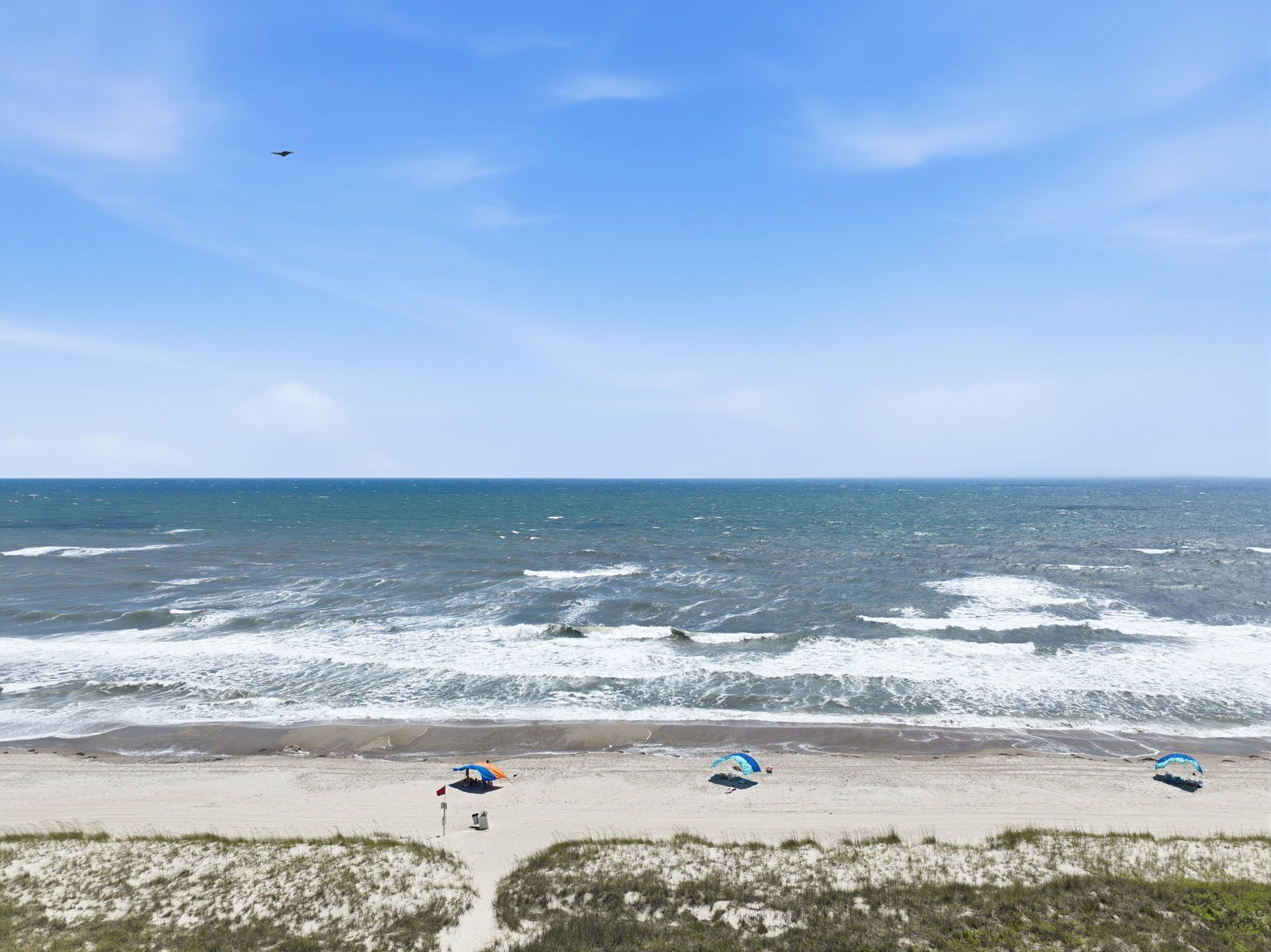 Une plage avec trois parasols, un drapeau et un oiseau dans le ciel.