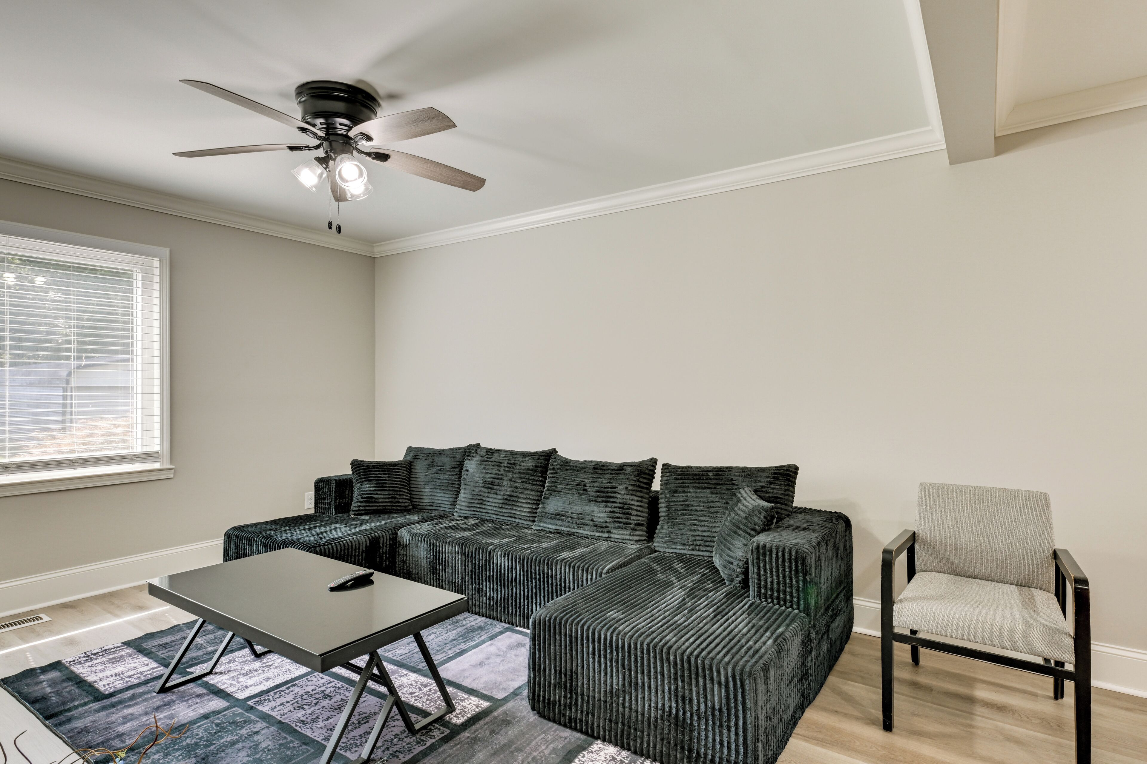 A living room with a sectional sofa, a small table, a chair, a ceiling fan, and a window with blinds.