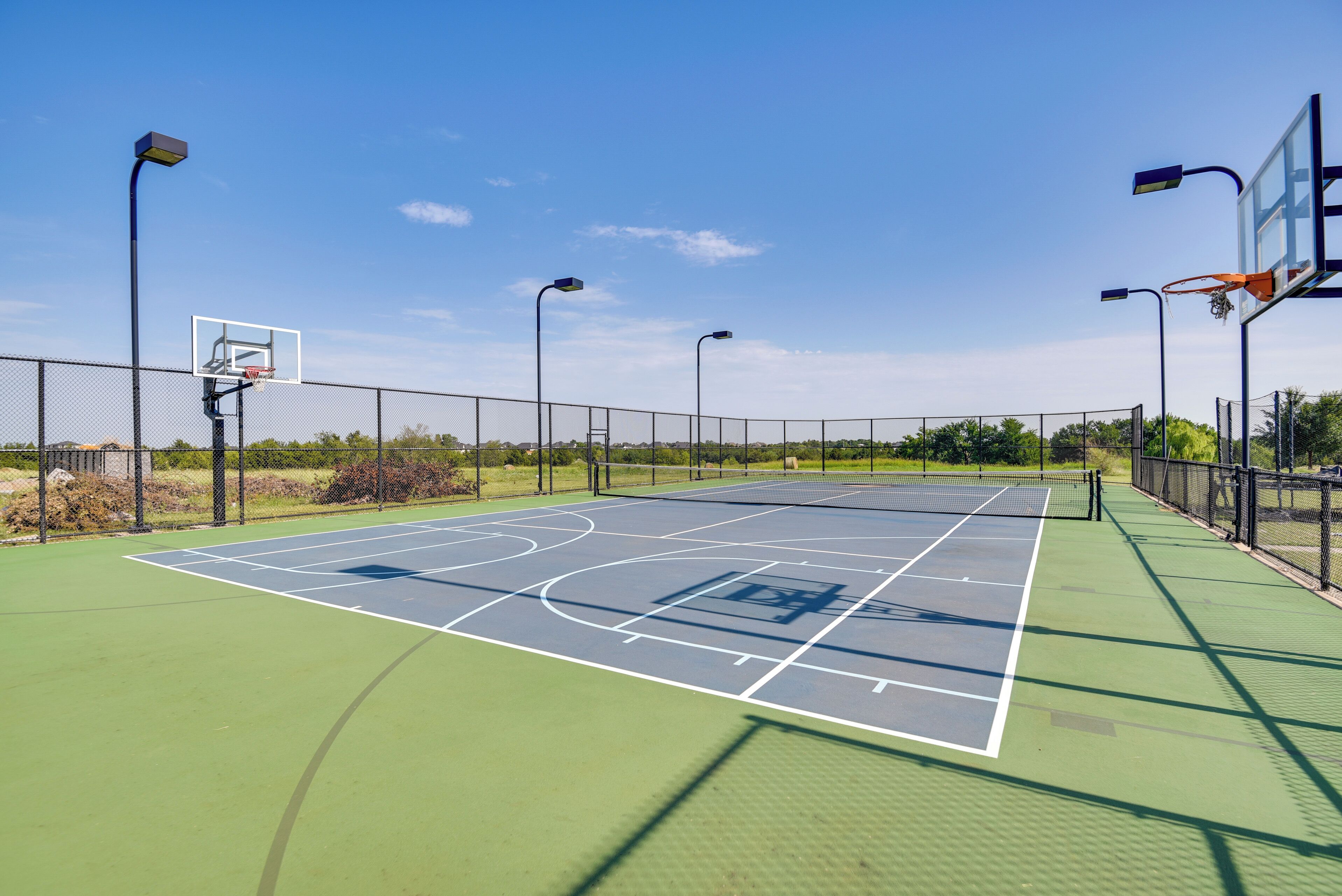 Un terrain de basket en plein air, doté d’une surface verte, entouré d’une clôture métallique et équipé d’un système d’éclairage suspendu.
