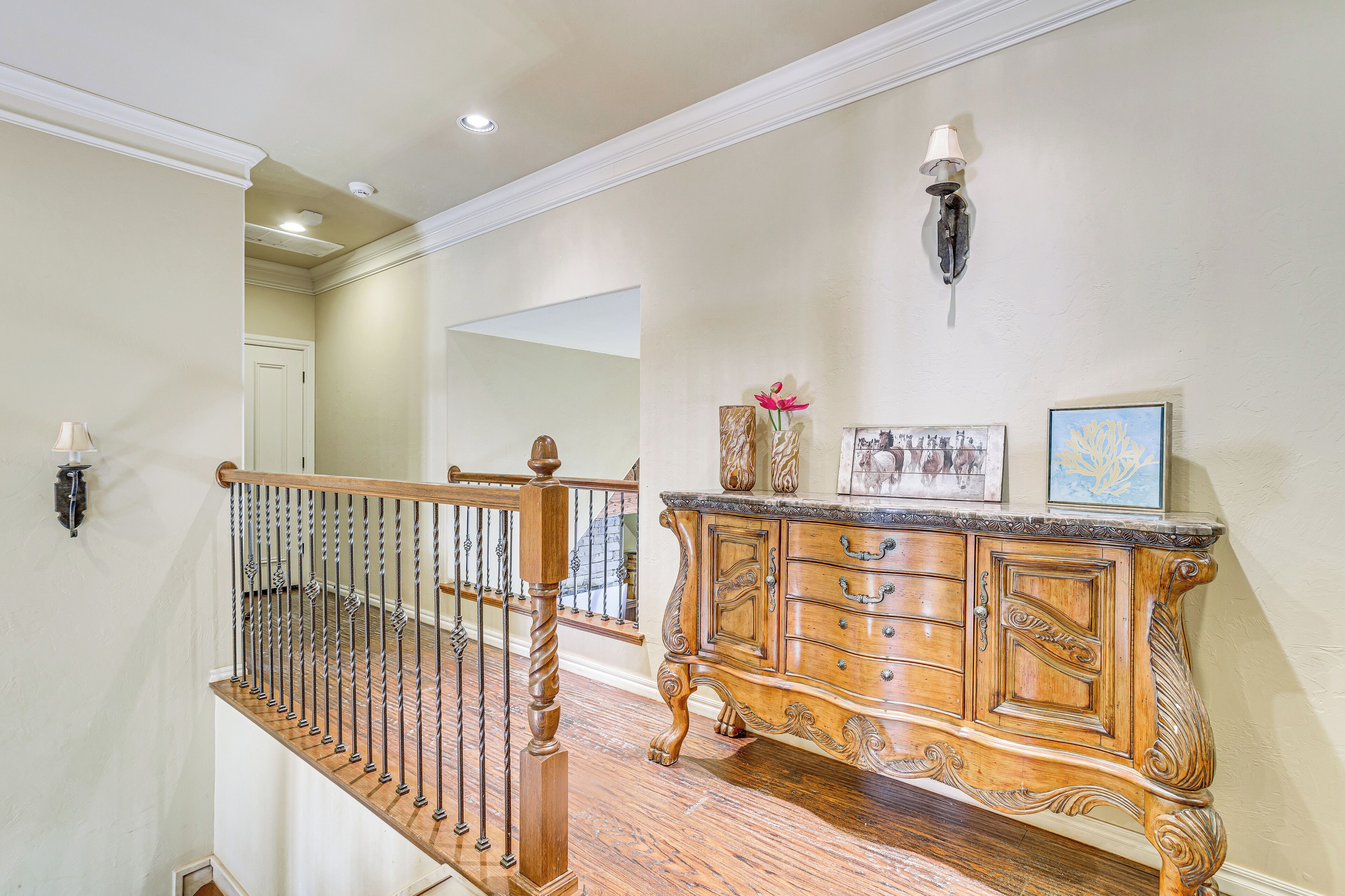 A staircase with a wooden handrail and metal balusters, leading to a landing with a marble-top console table, a framed picture, and a wall-mounted lamp.