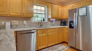 A kitchen with wooden cabinets, a stainless steel refrigerator, a dishwasher, and a window with a view of greenery.