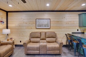 Una habitación acogedora con techo de madera, un cuadro enmarcado, un sofá, una mesita con una lámpara y una zona de bar con taburetes.