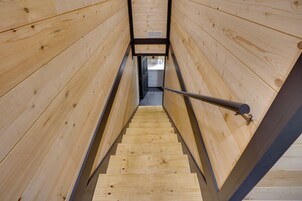 Una escalera interior de madera con barandillas de metal, que conduce a una habitación con una puerta y una rejilla de ventilación.