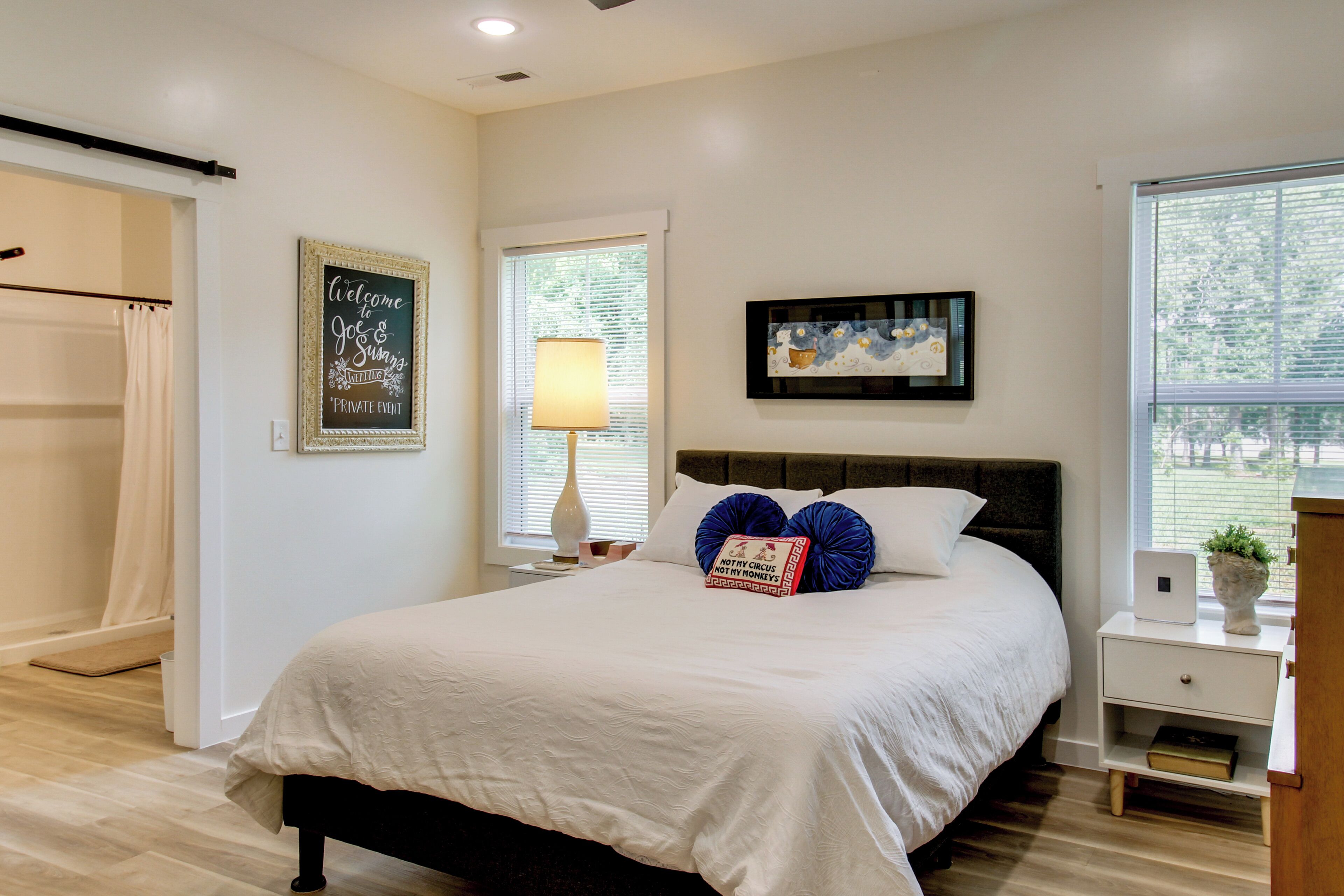 A bedroom with a bed, a bedside table, a lamp, a framed welcome sign, and a window with blinds.