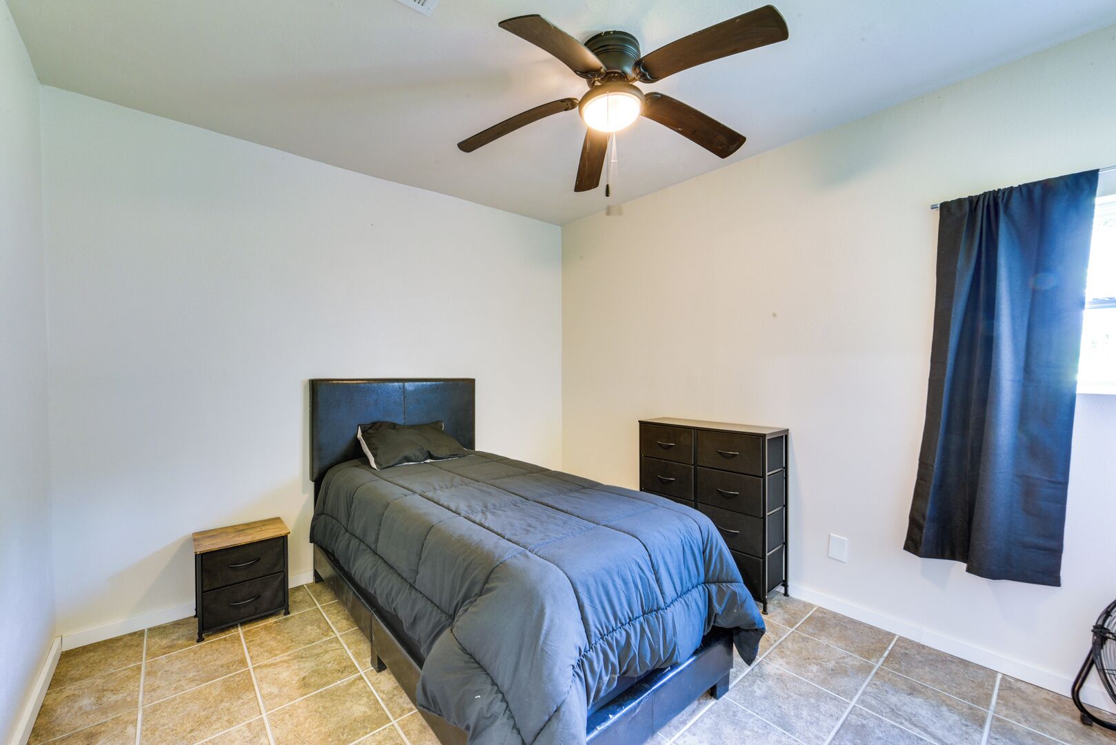 A bedroom with a bed, a nightstand, a dresser, a ceiling fan, and a window with curtains.