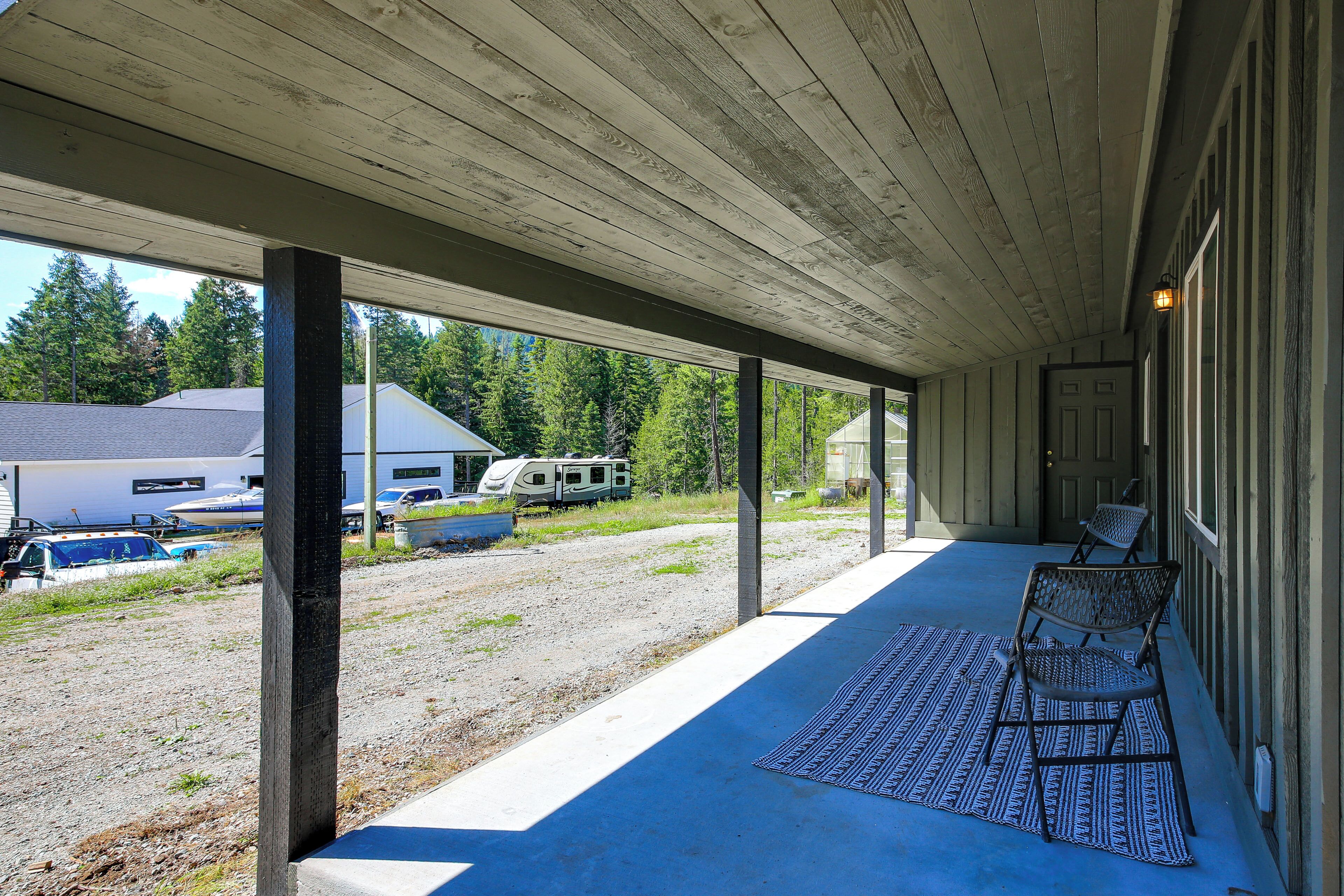A covered porch with a bench, a rug, and a view of a gravel area with parked vehicles and a house.