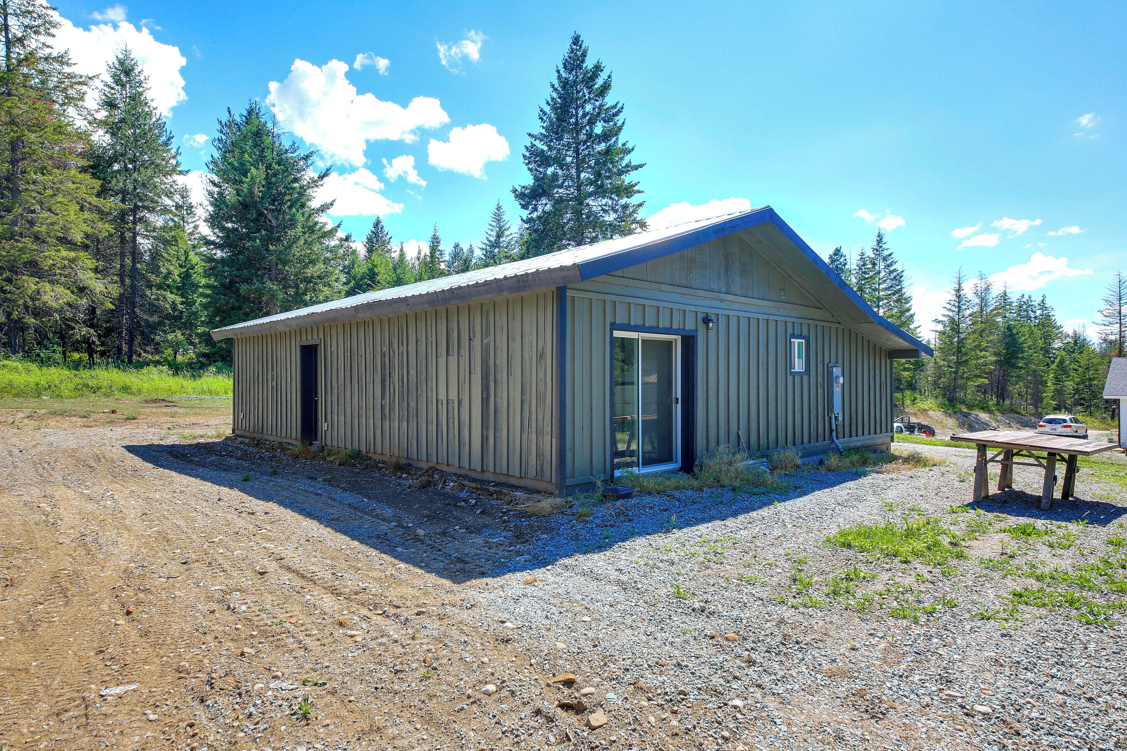 A single-story wooden building with a metal roof, a door, and a window, surrounded by gravel and trees.