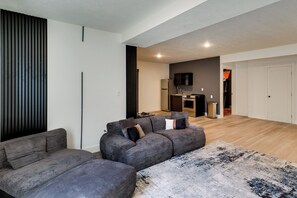 A modern living room with a sectional sofa, a flat-screen TV mounted on the wall, and a kitchen area in the background. - Quiet Tooele Retreat w/ Games + Fireplace (Tooele)