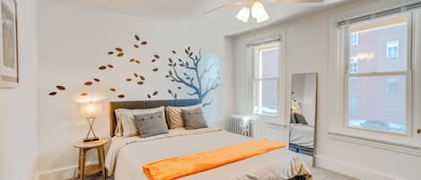Une chambre à coucher comprenant un lit, un ventilateur de plafond, une table de chevet, une lampe, une fenêtre et un élément de décoration murale.