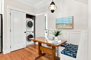 A laundry room with a washing machine, a table with a plant, and a bench with patterned cushions. - Seabrook Feelin TipSEA | Hot Tub, Dual Suites, 2BR | Sleeps 4, Lily Walk (Pacific Beach)