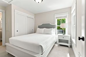 A bedroom with a bed, a nightstand, a window with blinds, and a door.