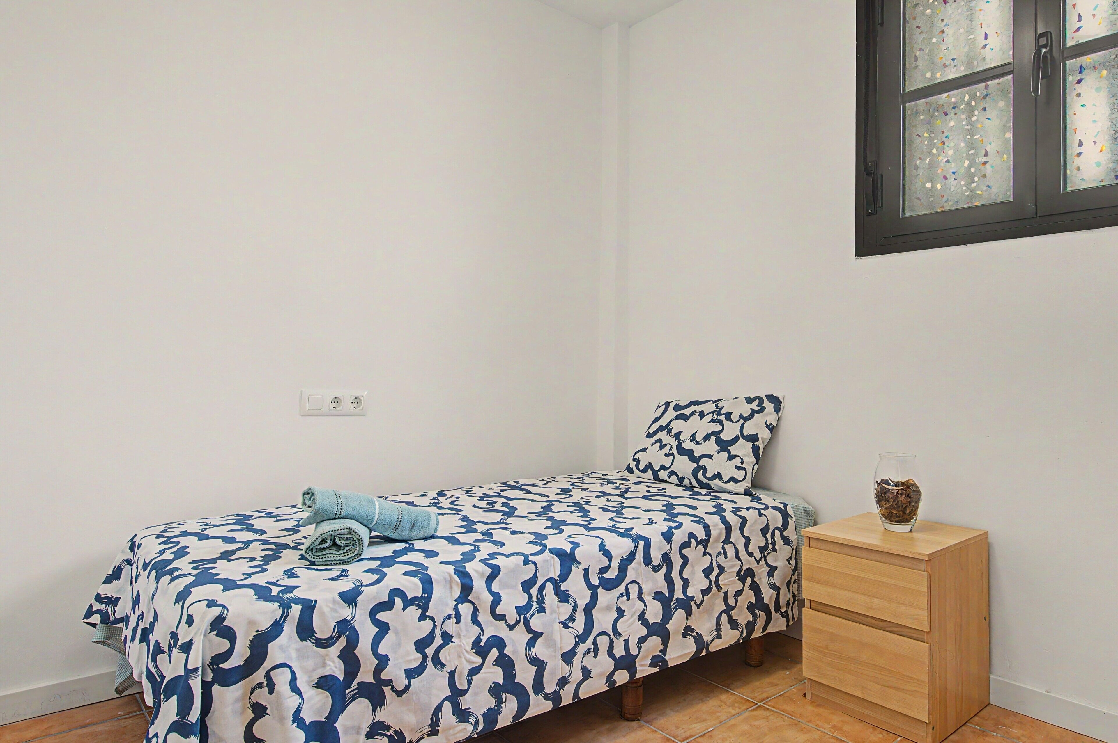 A single bed with a patterned bedspread, a bedside table with a vase, and a window with a view of the outside.