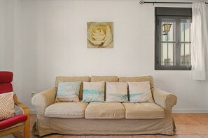 Ein beiges Sofa mit vier Kissen, ein roter Sessel, ein an der Wand hängendes Rosenbild und ein Fenster mit Vorhang.